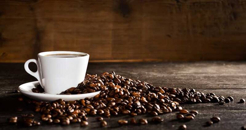 imagem de xícara branca com café, sobre um pirex, rodeada de diversos tipos de grãos de café