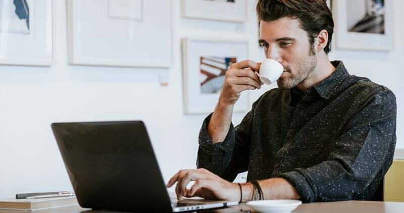 homem tomando café enquanto trabalha em frente a um notebook, representando a praticidade do comodato de máquina de café para empresas